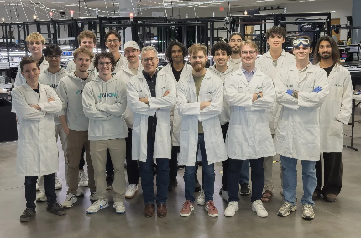 The Adom team in lab coats on the factory floor at 4400 Alliance Gateway in Fort Worth, with the workcell rows behind them